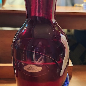 May include: A handcrafted, ruby red glass vase with a flared top and a blue spiral base. The vase has a smooth, glossy finish and a label that reads "LENKO HANDCRAFT". The vase is a decorative item.
