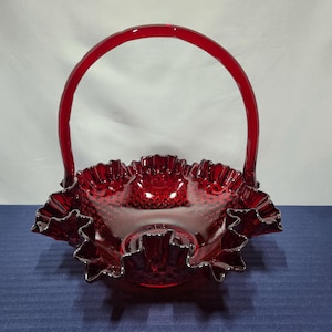 May include: A ruby red glass basket with a ruffled edge and a handle. The basket has a textured surface and a silver rim. The handle arches over the bowl. The basket is sitting on a blue surface.