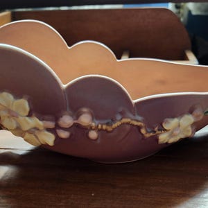 May include: A vintage, mauve-colored ceramic bowl with a scalloped rim and floral accents. The bowl features cream-colored flowers and brown branch-like handles. The interior is a light peach color.