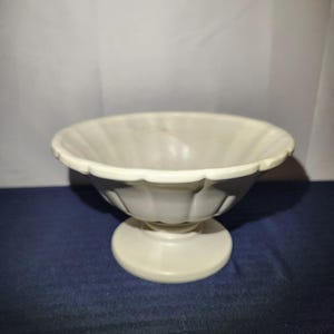 May include: A cream-colored, pedestal bowl with a scalloped rim. The bowl has a round base and a fluted design on the exterior. The bowl is made of ceramic or porcelain and is suitable for serving desserts or displaying decorative items.