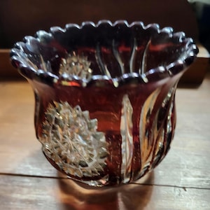 May include: A ruby-red glass votive candle holder with a scalloped rim. The sides feature a detailed, textured floral design. The glass has a glossy finish, reflecting light. The candle holder is designed to hold a small candle.
