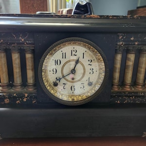 May include: A black mantel clock with a rectangular base and top, featuring four decorative columns with a marbled tan finish. The central circular clock face has a gold-toned border, black Arabic numerals, and two black hands on a white background.