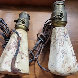 May include: Two vintage lamp bases with brass fixtures and cream-colored, marbled ceramic bodies. The bases have brown and blue veining and are connected by black and brown cords. The lamps are displayed against a wooden background.