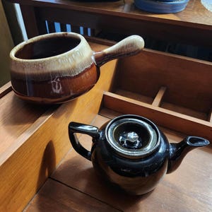May include: Two ceramic kitchen items on a wooden surface. A brown and tan handled soup bowl and a small black teapot with a lid. The soup bowl has a handle and a two-tone glaze. The teapot has a spout and a handle.