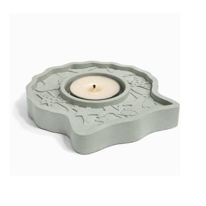 May include: A light gray concrete candle holder with a circular indentation for a tea light. The holder has a unique, organic shape with carved sea-themed designs. A lit white tea light candle sits in the center, casting a warm glow.