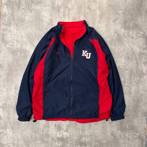 May include: Navy blue and red reversible jacket with a full zipper. The jacket features red side panels and a red interior. The left chest displays the white letters "KU". A small Nike logo is visible on the upper left chest.