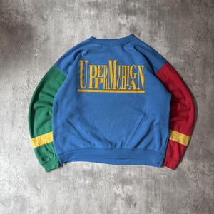 May include: A blue crewneck sweatshirt featuring the yellow text UPPER MICHIGAN across the chest. The left sleeve is green with a yellow stripe near the cuff, and the right sleeve is red with a yellow stripe near the cuff, creating a color-blocked design.