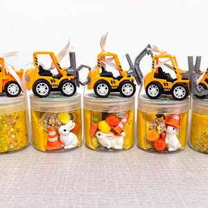 May include: Five clear jars filled with yellow slime and decorative elements, topped with toy construction vehicles. The jars contain glitter, small toys, and miniature construction cones and rabbits. The toy trucks are orange and black.