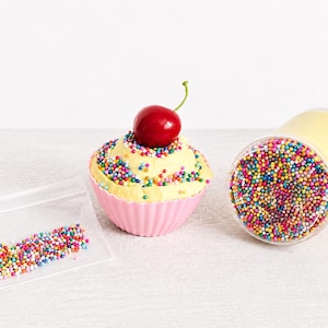 May include: A light yellow cupcake-shaped item in a pink fluted liner topped with multicolored sprinkles and a red cherry, a clear cylindrical container filled with yellow substance and topped with multicolored sprinkles, and a small plastic bag of sprinkles.