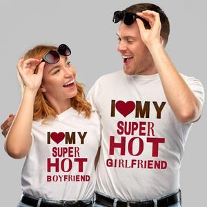 May include: Two people wearing white t-shirts with the text "I ❤️ MY SUPER HOT BOYFRIEND" and "I ❤️ MY SUPER HOT GIRLFRIEND" in maroon and brown. Both are wearing sunglasses and smiling.