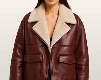 May include: A burgundy leather jacket with a cream-colored shearling collar and lining. The jacket features two front pockets and a zipper closure. The model is wearing a black top. The jacket is from Zain's Crafted.