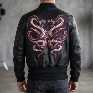 Könnte beinhalten: Eine schwarze Leder-Bomberjacke mit einer großen, detaillierten Stickerei auf der Rückseite. Das Design zeigt zwei ineinander verschlungene Schlangen in Dunkelviolett und Gold mit geöffneten Mäulern und sichtbaren Fangzähnen.