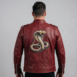 May include: A deep red leather jacket featuring a large, detailed graphic of a coiled cobra snake on the back. The jacket has a stand-up collar, shoulder epaulets, and silver-toned metal zips at the cuffs, showcasing a smooth, matte finish.
