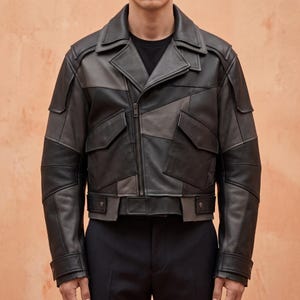 Puede incluir: Una chaqueta de cuero negra y gris carbón con cuello de solapa ancho, cierre de cremallera frontal asimétrico y dos grandes bolsillos con solapa en el pecho. La chaqueta incluye paneles estructurados en los hombros y un cinturón en la cintura.