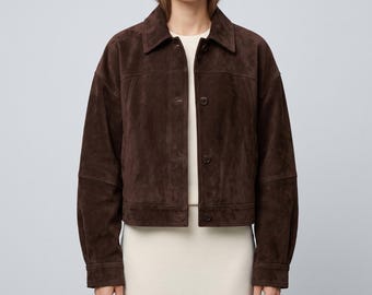 May include: A dark brown suede jacket featuring a classic collar, long sleeves with buttoned cuffs, and a front button closure. The jacket has a boxy, cropped silhouette with visible horizontal seam detailing across the chest and back.