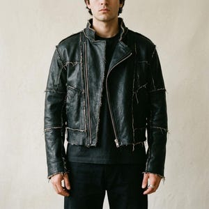 Puede incluir: Una chaqueta de cuero negro con acabado desgastado y costuras visibles, crudas y deshilachadas en toda la construcción. La chaqueta tiene un cierre de cremallera frontal asimétrico, cuello alzado y mangas largas sobre una camiseta negra.