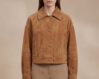 May include: A tan suede jacket featuring a pointed collar, long sleeves with buttoned cuffs, and a front button closure. The jacket has vertical seam detailing on the chest and two side pockets, paired with solid tan trousers in a neutral tone.