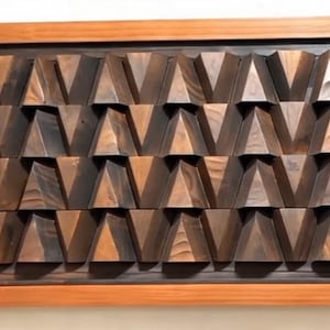 Puede incluir: Arte mural de madera con un patrón repetitivo de prismas triangulares. La obra de arte está enmarcada en un borde de madera natural, con los prismas que muestran diferentes tonos de marrón y una apariencia texturizada. Una pieza decorativa.
