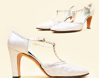 May include: A pair of silver glitter high-heeled shoes with a T-strap and braided detail. The shoes have a pointed toe and a block heel.
