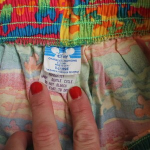 May include: A white tag with blue text attached to a colorful fabric. The tag reads "Character Fashions Disney Made in U.S.A. X-Large For care & other see reverse" and "Defenitiely Wash Gentle Cycle Do Not Bleach Flat to Dry".