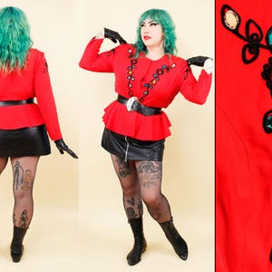 May include: A red long-sleeve jacket with decorative jewel accents, a black belt, and a black leather skirt. The jacket has a peplum design. The outfit is paired with black gloves and boots. The jacket is adorned with colorful jewels.
