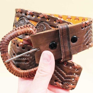 May include: Brown leather belt with intricate woven and cut-out designs. The belt features a large, rounded buckle and is secured with black snaps. The leather has a distressed appearance, adding to its vintage aesthetic. A stylish accessory for any outfit.