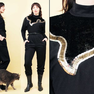 Op de afbeelding: Een zwarte jumpsuit met een gouden en zwart paillettenontwerp aan de voorkant. De jumpsuit heeft een hoge hals en lange mouwen. Het model draagt zwarte laarzen.