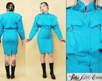 May include: A turquoise two-piece outfit, featuring a long-sleeved top with shoulder pads and a pencil skirt. The top has a high collar, decorative metal rings, and pockets. The skirt has a high waist and a slit in the back. The outfit is from Saks Fifth Avenue.