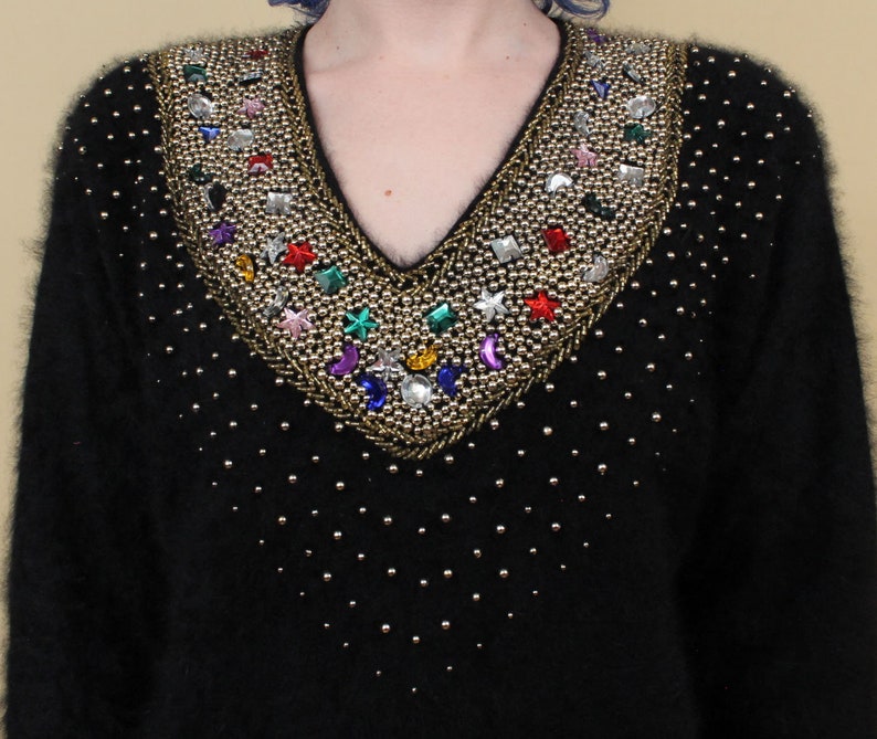 May include: A black fuzzy sweater with a V-neck and a beaded design. The beads are gold, silver, and multi-colored and form a pattern of stars, moons, and geometric shapes.