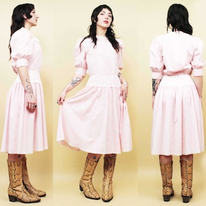 Könnte beinhalten: Ein rosa Kleid mit weißer Spitzenborte und einem gerafften Rock. Das Kleid hat ein tailliertes Oberteil und kurze Ärmel. Das Model trägt braune Schlangenlederstiefel.