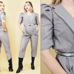 Op de afbeelding: Een grijze jumpsuit met een gekraagde, knoopsluiting en wijde pijpen. De jumpsuit heeft een riem en pofmouwen. Het model draagt zwarte laarzen.