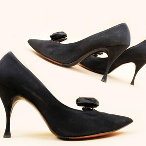Op de afbeelding: Een paar zwarte pumps met hoge hakken, spitse neuzen en een kleine strik. De slanke hakken en oranje zolen geven een vintage uitstraling. Klassieke damesschoenen.