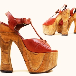 May include: A pair of vintage platform sandals with a wooden base and a red leather upper. The sandals have a T-strap design and a buckle closure. The platform measures approximately 4 inches (10 cm) high.