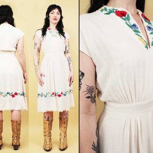 May include: A white vintage dress with a V-neck and short sleeves. The dress features colourful embroidered flowers around the neckline and hem. The dress is knee-length and has a fitted bodice and a flowing skirt.