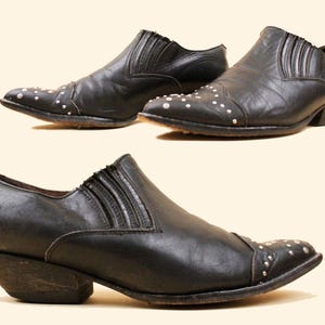 May include: A pair of black leather slip-on shoes featuring a pointed toe with silver-tone metal stud embellishments, a low stacked heel, and vertical stitched pleat detailing along the side panels of the upper construction.