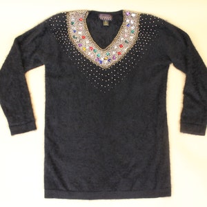 May include: Black fuzzy sweater with a v-neck and a large jeweled embellishment around the neckline. The jewels are gold, red, blue, green, and clear.