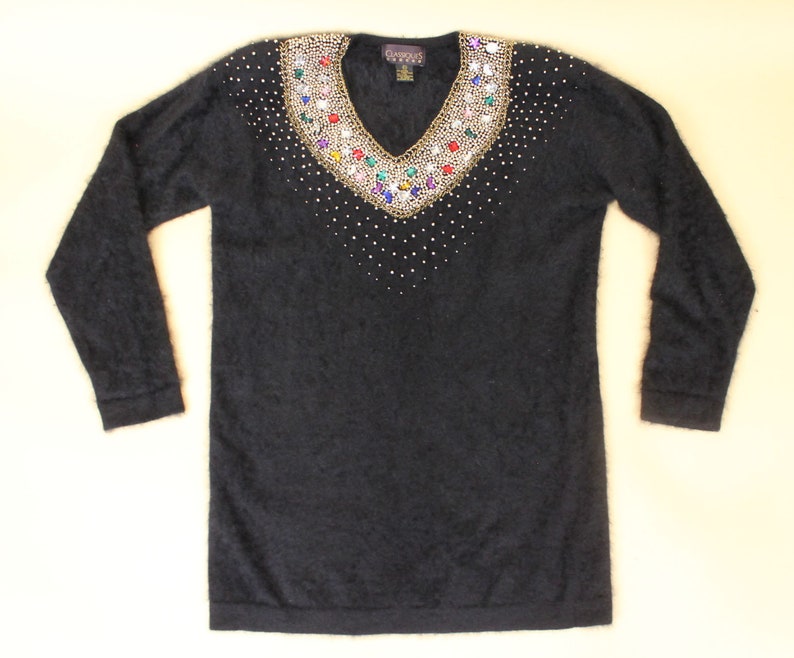 May include: Black fuzzy sweater with a v-neck and a large jeweled embellishment around the neckline. The jewels are gold, red, blue, green, and clear.
