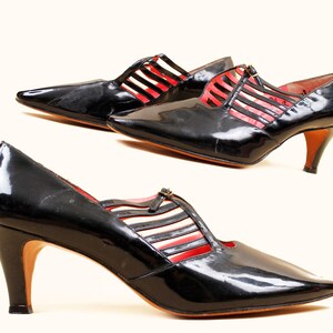 May include: Pair of black patent leather pumps with pointed toes and small heels. The shoes feature a cut-out design with red and white accents. The heels are approximately 2 inches high.