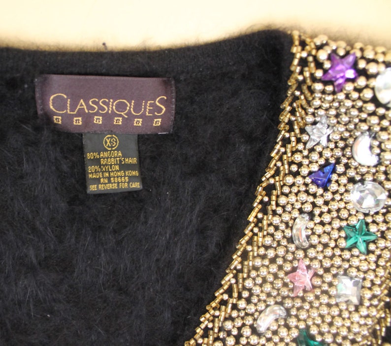 May include: Black angora rabbit hair and nylon blend cardigan with a beaded neckline featuring gold beads, rhinestones, and colorful stars. The label reads "CLASSIQUES" and "80% Angora Rabbit's Hair 20% Nylon Made in Hong Kong RN 58665 See Reverse for Care".