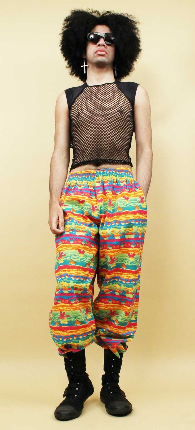 May include: A person wearing a black mesh tank top, colorful patterned pants, and black boots. The pants have a bright, multi-colored pattern with a geometric design.