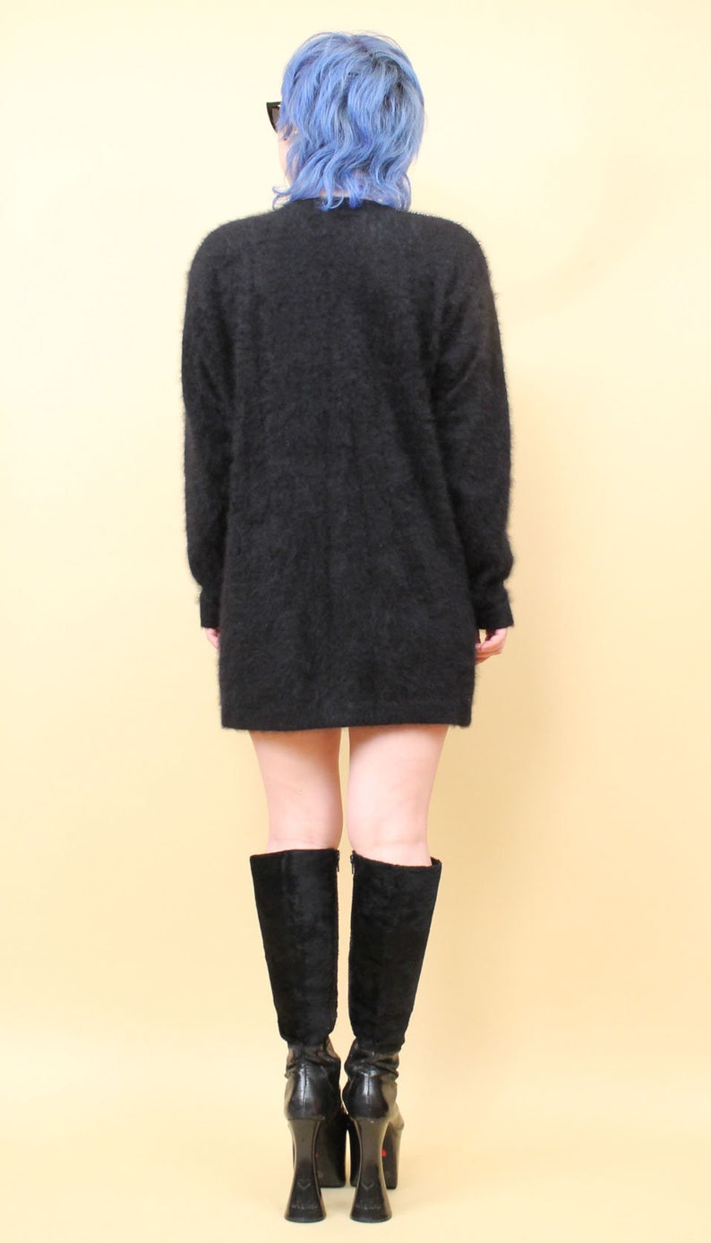 May include: A black fuzzy sweater with long sleeves and a loose fit. The sweater is worn over a pair of black knee-high boots with a platform heel.
