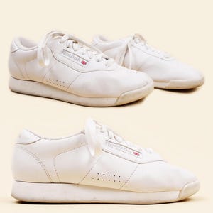 Op de afbeelding: Een paar witte Reebok sneakers met veters, op een lichtbeige achtergrond. De sneakers hebben een laag profiel, met het Reebok logo aan de zijkant. De schoenen lijken gedragen, met wat verkleuring op de zolen.