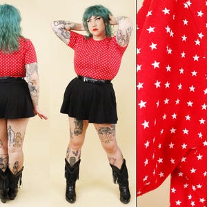 Puede incluir: Una camiseta roja de manga corta con un patrón repetido de estrellas blancas, combinada con una minifalda negra y un cinturón negro. La camiseta tiene cuello redondo y un corte ajustado, mientras que la falda es negra con un dobladillo acampanado.