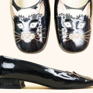 May include: A pair of black patent leather shoes with a black cat face embroidered on the toe. The shoes have a low heel and a pointed toe.