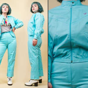 Op de afbeelding: Een turquoise blauwe trainingspak met een ritsjack en bijpassende broek. Het jack heeft een opstaande kraag en een klein logo aan de rechterkant van de borst. De broek heeft een gezoomde zoom.