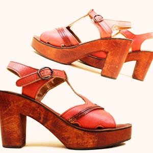 May include: A pair of vintage platform sandals. The sandals feature a reddish-orange leather upper with a T-strap design and a buckle closure. They have a wooden platform sole and a chunky wooden heel.