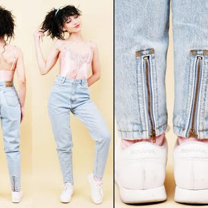 May include: Light wash denim jeans with ankle zippers. The jeans have a high waist and a classic fit. The model is wearing a pink camisole and white sneakers. The jeans are a vintage style.