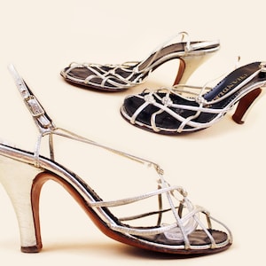 May include: Silver high-heeled sandals with a cage-like design. The sandals have a metallic silver finish, a strap around the ankle, and a brown heel. The shoes are displayed on a light beige background. The heels are approximately 3 inches high.