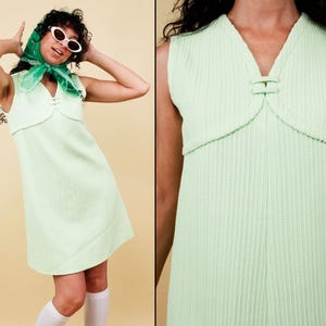 Könnte beinhalten: Hellgrünes Vintage-Kleid mit V-Ausschnitt und dekorativem Detail an der Taille. Das Kleid hat einen strukturierten, gerippten Stoff und ein ärmelloses Design. Das Model trägt eine weiße Sonnenbrille und einen grünen Schal.