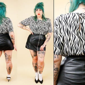May include: A short-sleeved blouse featuring a black and white zebra stripe pattern with a round neckline and a back button closure, paired with a black mini skirt that has a central vertical silver zipper running down the front.